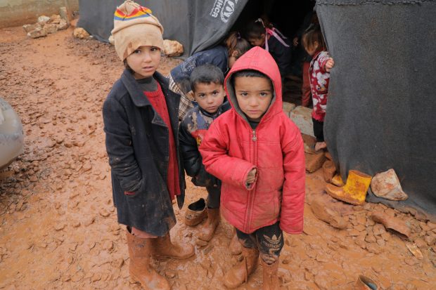 Displaced Syrian children stand outside in the cold mud outside their tent.